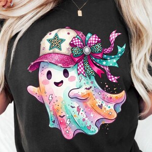 Puede incluir: Una camiseta negra con un gráfico colorido de un fantasma que lleva una gorra de béisbol rosa brillante con una estrella verde azulada. El fantasma está adornado con un gran lazo estampado en rosa y verde azulado, murciélagos, corazones y rayos.