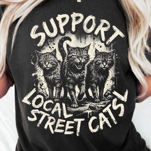 Puede incluir: Una camiseta negra con un estampado gráfico de tres gatos en el centro. El texto "SUPPORT LOCAL STREET CATS" está arqueado sobre y debajo de los gatos en una fuente blanca desgastada. El diseño tiene una estética de ilustración estilo tinta.
