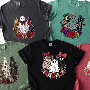 Puede incluir: Cinco camisetas de manga corta y cuello redondo en gris carbón, verde azulado, verde salvia, negro y granate. Cada camiseta presenta un estampado gráfico de tres fantasmas con lazos estampados sobre un grupo de rosas y flores.