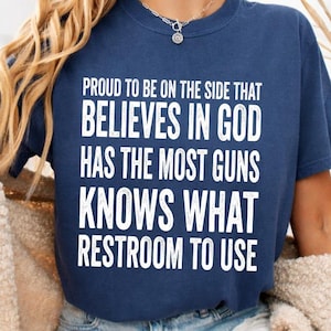 May include: A navy blue short-sleeved t-shirt featuring white text that reads: PROUD TO BE ON THE SIDE THAT BELIEVES IN GOD HAS THE MOST GUNS KNOWS WHAT RESTROOM TO USE. The shirt has a crew neckline and is worn with light blue denim shorts.