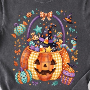 Puede incluir: Una camiseta gris carbón con un gráfico de una cesta de dulces en forma de calabaza llena de caramelos, estrellas y un sombrero de bruja. El diseño incluye una calabaza a cuadros, un cupcake, un gusano y pequeños lazos en naranja, morado y azul.