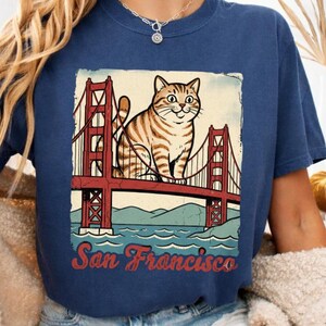 Puede incluir: Una camiseta azul marino con un estampado gráfico de un gato atigrado marrón sentado sobre el puente Golden Gate. Debajo del puente, el texto San Francisco está impreso en una fuente cursiva roja. La camiseta tiene cuello redondo y mangas cortas.