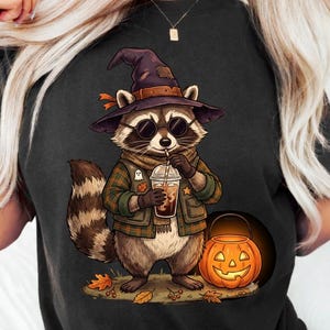 Puede incluir: Una camiseta negra con el gráfico de un mapache que lleva un sombrero de bruja y gafas de sol, vestido con una camisa de cuadros y un chaleco. El mapache sostiene un vaso de plástico con pajita junto a una calabaza tallada de color naranja.