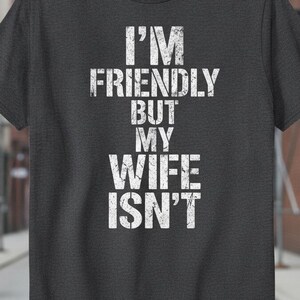 Puede incluir: Una camiseta de manga corta en color gris carbón oscuro con el texto blanco de estilo desgastado I'M FRIENDLY BUT MY WIFE ISN'T impreso en el centro del pecho. La prenda tiene cuello redondo y un corte estándar con dobladillo recto.