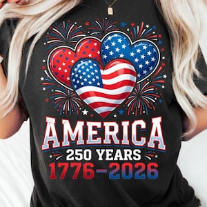 Puede incluir: Una camiseta negra con un gráfico de tres corazones decorados con estrellas y rayas rojas, blancas y azules, rodeados de fuegos artificiales. El texto AMERICA 250 YEARS 1776-2026 aparece en letras blancas, rojas y azules.
