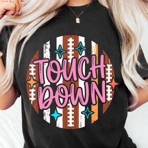 Puede incluir: Una camiseta negra de manga corta con un gráfico circular de rayas verticales marrones y blancas que recuerda a un balón de fútbol americano. El texto "TOUCH DOWN" aparece en grandes letras rosas, decorado con pequeñas estrellas azules y naranjas.
