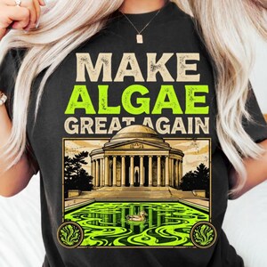 Könnte beinhalten: Ein schwarzes T-Shirt mit dem Text "MAKE ALGAE GREAT AGAIN" in Beige und leuchtendem Grün. Unter dem Text befindet sich eine grafische Illustration eines neoklassizistischen Gebäudes mit Säulen hinter einem Teich voller leuchtend grüner Algen und einer Ente.