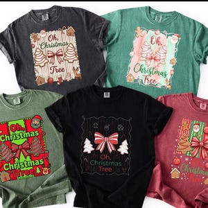 Puede incluir: Cinco camisetas de manga corta en gris carbón, verde salvia, verde oliva, negro y granate. Cada camiseta presenta un gráfico central con el texto Oh, Christmas Tree, rodeado de ilustraciones de galletas, lazos y adornos navideños.