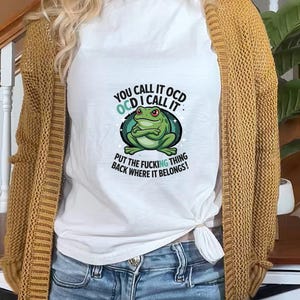 Puede incluir: Camiseta blanca con un gráfico de rana verde y el texto "YOU CALL IT OCD, OCD I CALL IT, PUT THE FUCKING THING BACK WHERE IT BELONGS!". La camiseta se usa con vaqueros azules y una rebeca amarillo mostaza.