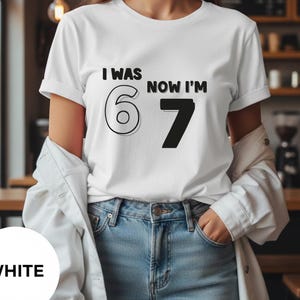 Puede incluir: Camiseta blanca con el texto negro "I WAS 6 NOW I'M 7". El número 6 está delineado y el número 7 es negro. La camiseta se usa con vaqueros azules y una chaqueta blanca.