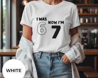 May include: White t-shirt with the text "I WAS 6 NOW I'M 7" in black. The number 6 is outlined, and the number 7 is solid black. The shirt is worn with blue jeans and a white jacket.