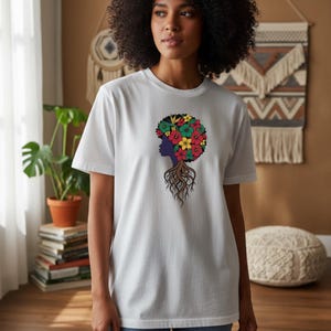 Puede incluir: Camiseta blanca con un gráfico de la silueta de una mujer con afro floral y raíces. El diseño utiliza rojo, amarillo, verde y morado. La camiseta es de cuello redondo.