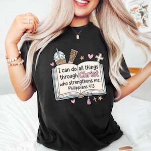 Puede incluir: Una camiseta negra con un gráfico de un libro abierto con el texto 'I can do all things through Christ who strengthens me. Philippians 4:13'. El diseño incluye ilustraciones de una taza de café, un tubo con estampado de leopardo, corazones y estrellas.