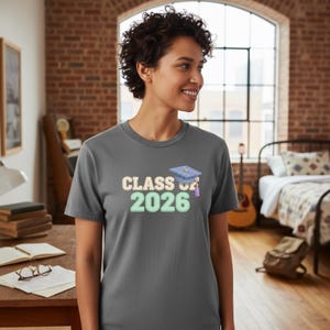 Puede incluir: Una camiseta gris con el texto "CLASS OF 2026" en colores pastel y un gráfico de birrete. La camiseta la lleva una persona con el pelo corto y rizado.