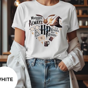Puede incluir: Camiseta blanca con un diseño en forma de corazón con el texto "Muggles Always HP" y otros elementos de una popular serie de fantasía. El diseño incluye una snitch dorada, gafas, un sombrero seleccionador y la frase "It's Leviosa, not Leviosa."