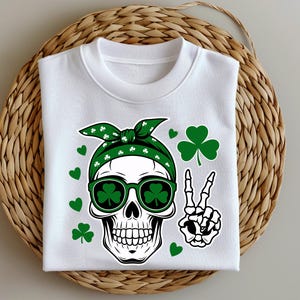 Puede incluir: Sudadera blanca con un diseño del Día de San Patricio. El gráfico incluye una calavera con una bandana verde, gafas de sol con tréboles y un signo de la paz de huesos. Tréboles y corazones verdes dispersos.