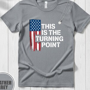 Puede incluir: Camiseta gris jaspeado con una bandera americana desgastada y el texto blanco "THIS IS THE TURNING POINT". La camiseta tiene cuello redondo y mangas cortas. Se ve un collar plateado con un colgante redondo.