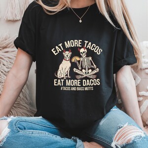 Puede incluir: Camiseta negra con un gráfico blanco de un esqueleto y un perro sosteniendo tacos. El texto en la camiseta dice "Eat More Tacos Eat More Dacos #Tacos and Biass Mutts".