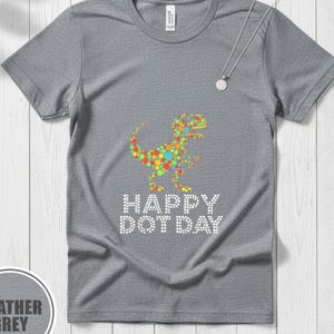 Puede incluir: Camiseta gris jaspeado con un diseño de dinosaurio colorido con puntos. El texto "HAPPY DOT DAY" está impreso debajo del dinosaurio en puntos blancos. También se ve un collar plateado con un colgante redondo.