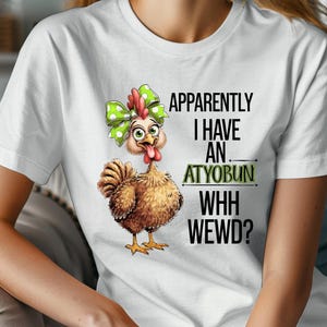 Puede incluir: Camiseta blanca con un pollo marrón con un lazo verde y blanco de lunares y el texto "Apparently I have an atyobun whh wewd?"