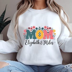 Puede incluir: Sudadera blanca con un diseño floral y la palabra "MOM" en letras de colores. El texto "Elizabeth & Miles" está impreso debajo de la palabra "MOM".