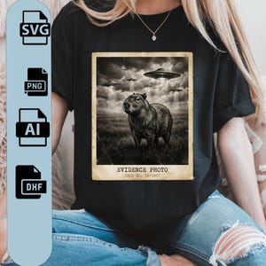 Puede incluir: Camiseta negra con una imagen en tonos sepia de un carpincho bajo OVNIs. La imagen está etiquetada como "EVIDENCE PHOTO" y "CASE NO. 7A-1967". La camiseta también tiene iconos de tipo de archivo SVG, PNG, AI y DHF.