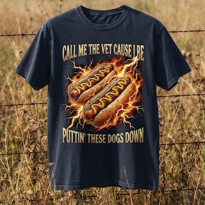 Puede incluir: Una camiseta de manga corta azul marino oscuro con un gráfico de dos perritos calientes con mostaza amarilla, rodeados de llamas naranjas y rayos amarillos. El texto dice: CALL ME THE VET CAUSE I BE PUTTIN' THESE DOGS DOWN en letras doradas.