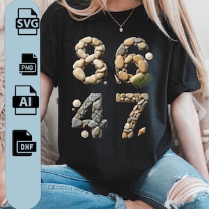 Puede incluir: Camiseta negra con los números 8647, cada número hecho de pequeñas piedras lisas. El diseño incluye una pequeña hoja de palma y conchas marinas. La camiseta se combina con vaqueros azules.