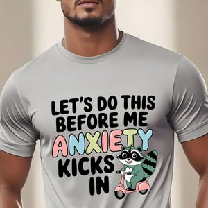 Puede incluir: Una camiseta gris con el texto "Let's do this before me anxiety kicks in" en negro. Un mapache de dibujos animados conduciendo un scooter está por encima del texto. El mapache es verde, negro y blanco con orejas rosas y una nariz azul.