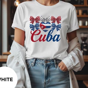 Puede incluir: Camiseta blanca con la palabra "Cuba" en escritura roja y azul. Encima de la palabra hay un corazón con la bandera cubana y varios lazos en rojo, blanco y azul. La camiseta está hecha de un material suave.