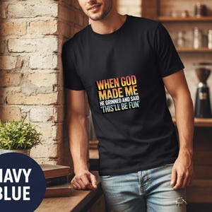 Puede incluir: Camiseta negra con el texto "WHEN GOD MADE ME HE GRINNED AND SAID 'THIS'LL BE FUN'" en un degradado colorido. La camiseta se combina con unos vaqueros azules. Un círculo azul marino con las palabras "NAVY BLUE" está en la esquina inferior izquierda.