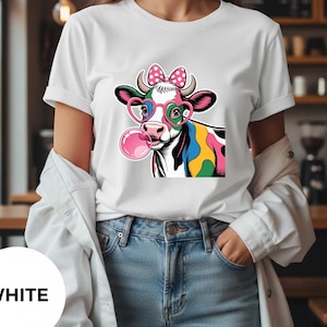 Puede incluir: Camiseta blanca con una vaca de dibujos animados. La vaca tiene gafas en forma de corazón, un lazo rosa y una burbuja de chicle. La vaca tiene manchas azules, amarillas y rosas. La palabra "WHITE" está en un círculo blanco en la parte inferior.