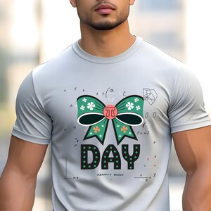 Puede incluir: Camiseta gris claro con un gran diseño de lazo verde con detalles blancos y naranjas. La palabra "DAY" está impresa en negro con lunares verdes. La camiseta también incluye símbolos matemáticos y el texto "HAPPY F. BUOLK".