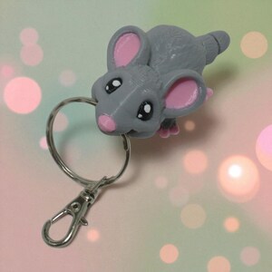 以下が含まれることがあります： ピンクの耳、鼻、足が付いた灰色のマウスキーホルダー。マウスは大きな黒い目を持ち、シルバーのキーリングと留め金に取り付けられています。キュートなアクセサリーです。
