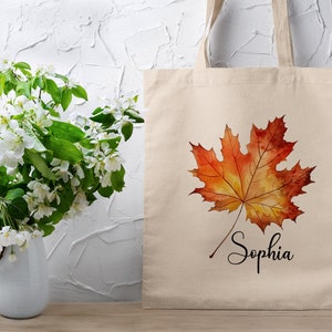 May include: A beige tote bag featuring a watercolour painting of an orange and yellow maple leaf. The bag is personalised with the name "Sophia" written in black cursive below the leaf.