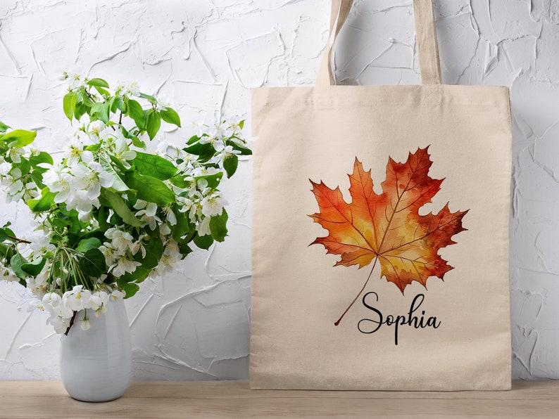 May include: A beige tote bag featuring a watercolour painting of an orange and yellow maple leaf. The bag is personalised with the name "Sophia" written in black cursive below the leaf.