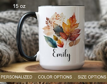May include: White ceramic mug with a black handle. The mug features a watercolor design of autumn leaves in shades of orange, yellow, red, and green. The name "Emily" is written in black cursive lettering below the leaves. The mug is 15 ounces.