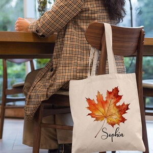 May include: A white canvas tote bag featuring a watercolour illustration of an orange and yellow maple leaf. The bag is personalised with the name "Sophia" written in cursive below the leaf.