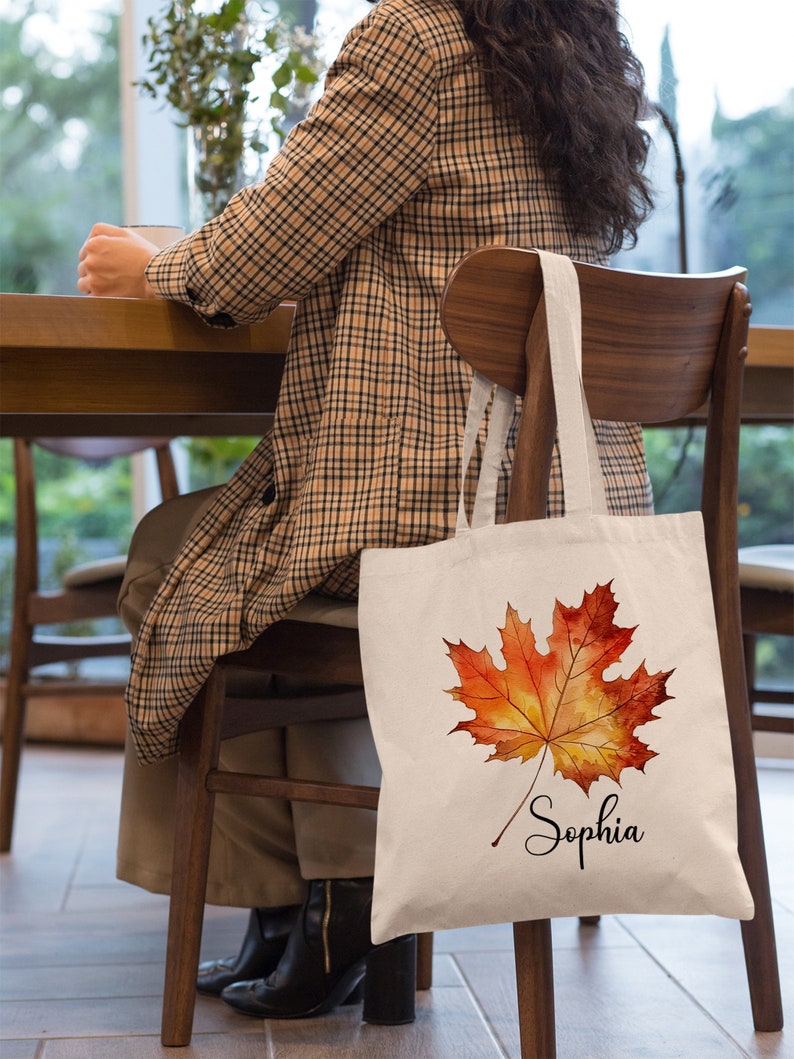 May include: A white canvas tote bag featuring a watercolour illustration of an orange and yellow maple leaf. The bag is personalised with the name "Sophia" written in cursive below the leaf.