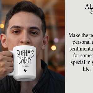 Peut inclure: Une tasse &agrave; caf&eacute; en c&eacute;ramique blanche avec du texte noir qui dit "Sophia's Daddy Est. 2025" et deux petites empreintes de pas.