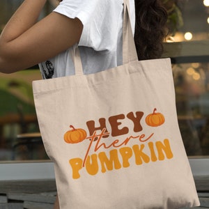 May include: A beige tote bag with the text "Hey there pumpkin" in orange and brown letters. Two small orange pumpkins are on either side of the text.