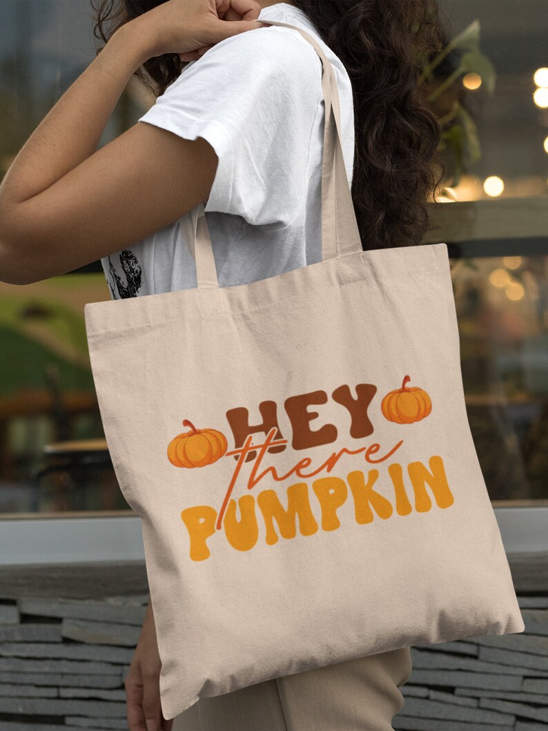May include: A beige tote bag with the text "Hey there pumpkin" in orange and brown letters. Two small orange pumpkins are on either side of the text.