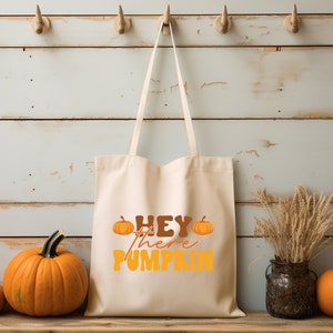 May include: A white canvas tote bag with the text "Hey there pumpkin" in orange and brown lettering. Two small orange pumpkins are on either side of the text. The bag is hanging on hooks against a white wood background. Three pumpkins are in the foreground.