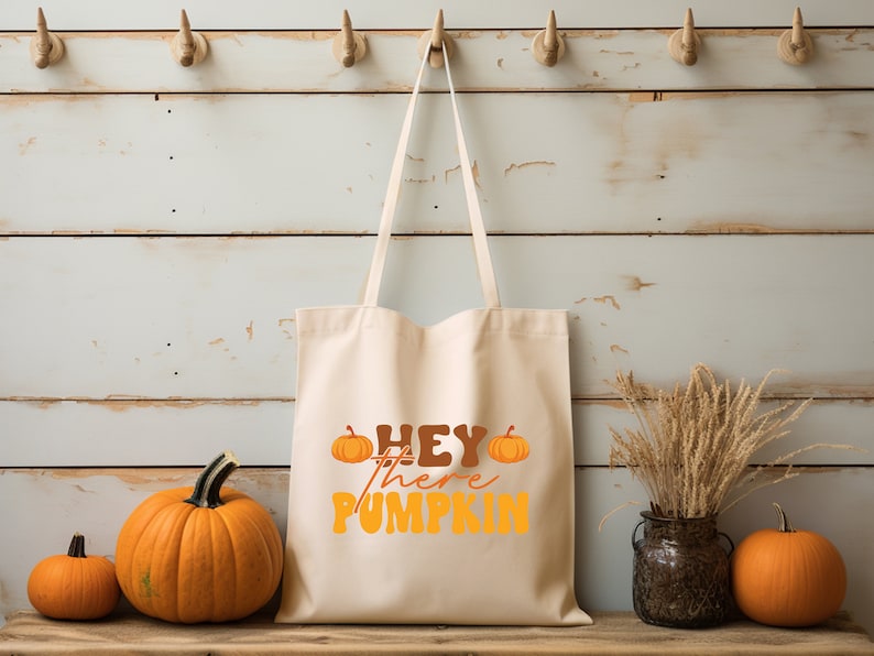 May include: A white canvas tote bag with the text "Hey there pumpkin" in orange and brown lettering. Two small orange pumpkins are on either side of the text. The bag is hanging on hooks against a white wood background. Three pumpkins are in the foreground.