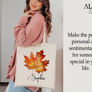 May include: A white tote bag featuring a watercolour illustration of an autumn leaf in shades of orange and yellow. The name "Sophia" is written in cursive below the leaf.