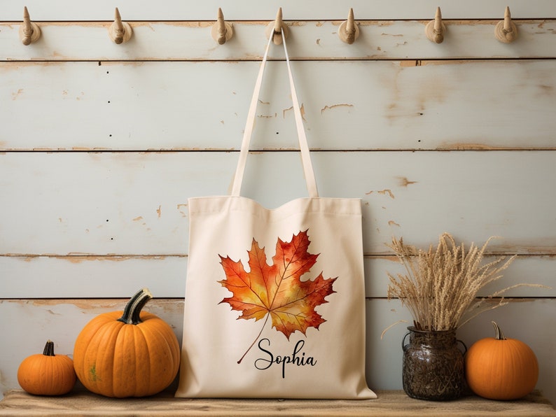 May include: A beige canvas tote bag featuring a watercolour-style orange and yellow maple leaf design. The bag has a black handle and the name "Sophia" is written in cursive below the leaf.
