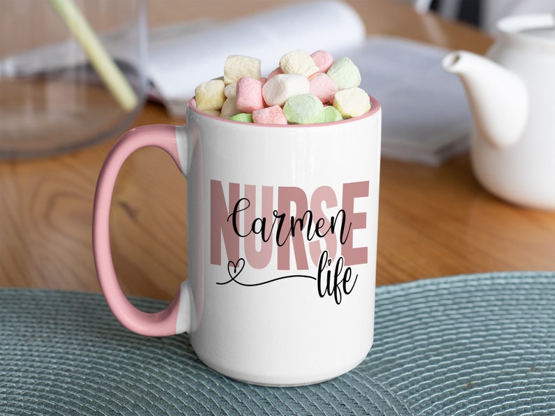 Puede incluir: Taza de cer&aacute;mica blanca con borde y asa de color rosa. La taza tiene el texto "NURSE carmen life" impreso en letras rosa y negras. La taza est&aacute; llena de malvaviscos.