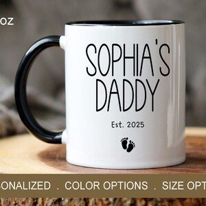 Peut inclure: Une tasse en c&eacute;ramique blanche avec une anse et un bord noirs. La tasse porte une inscription noire "Sophia's Daddy Est. 2025" et un petit graphique de pied.