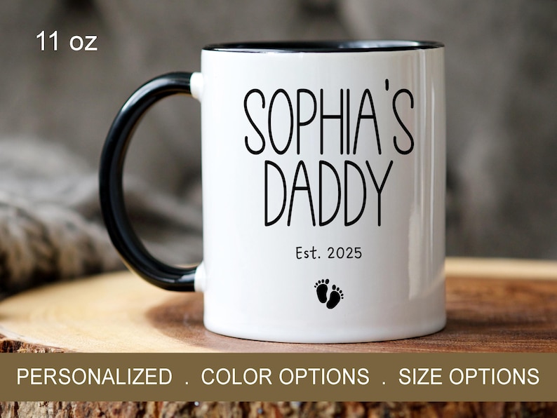 Peut inclure: Une tasse en c&eacute;ramique blanche avec une anse et un bord noirs. La tasse porte une inscription noire "Sophia's Daddy Est. 2025" et un petit graphique de pied.