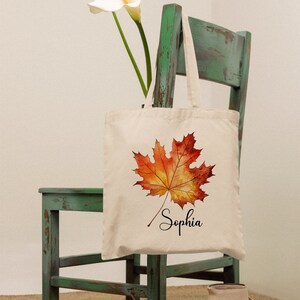 May include: A white canvas tote bag featuring a watercolour illustration of an orange and yellow maple leaf. The bag is inscribed with the name "Sophia" in cursive below the leaf. The bag is hanging on a green wooden chair.
